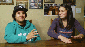 Dani smiles and raps while Athena looks at her, smiles, and keeps the beat on the dining room table with her hands.