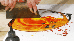 Kris Haas's hands holding a palette knife, scraping it along the paint to create a fan-like circle.