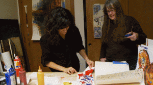 Kris Haas watches in her studio as Cheryl Green dabs an abstract painting with a small square of paper covered in spilled red paint.
