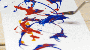 A small palette knife held above white paper. On the paper are swirls, streaks, and blobs of bright cobalt, red, and yellow paint.