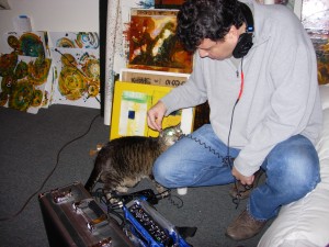 Alex Kitty tastes some cable as Esteban crouches on the floor and sets up to record at Kris Haas's studio. He is surrounded by paintings.