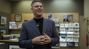 Jeff Black wears a suit and smiles in front of a display of art and frames on the wall.