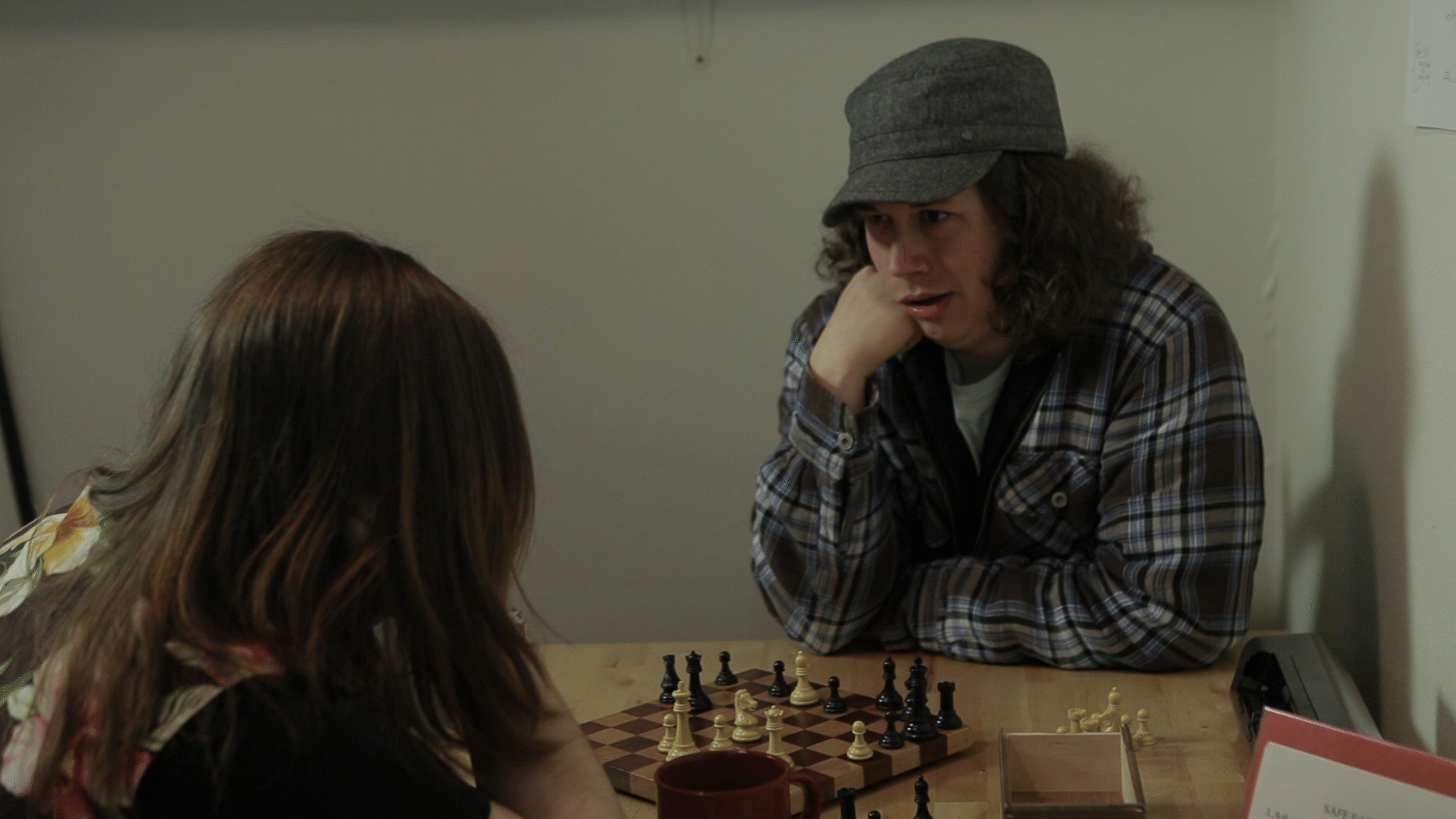 A young white man and woman play chess. Both people have their right elbows resting on the table with their heads resting on their own right hands.