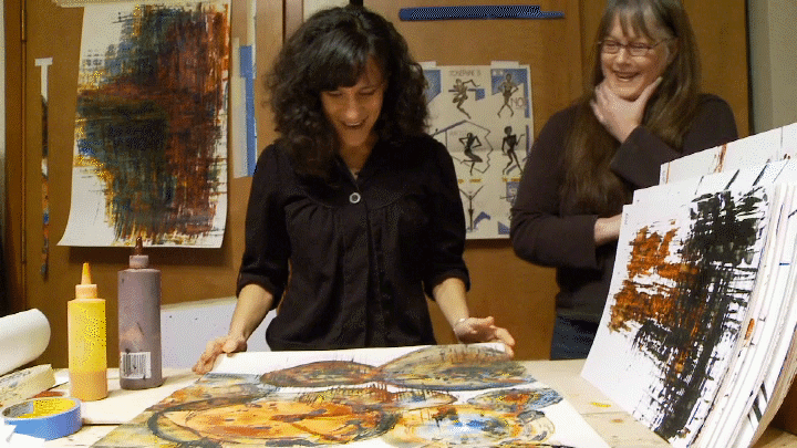 Two women in black shirts in an art studio with abstract paintings on the table and wall. Kris Haas stands back, smiling. Cheryl Green holds a large painting with circles in rust, yellow, blue, and green and looks surprised.