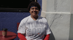 A smiling young Black woman with short hair and a blue sweat band sits outside. She has a white t-shirt with "I was born to ball" on it and iPod earbuds around her neck. 
