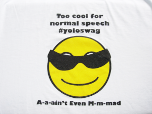 Screen-print on a t-shirt of "Too cool for normal speech." Below is a yellow smiley face icon wearing sunglasses and "A-a-ain't Even M-m-mad."