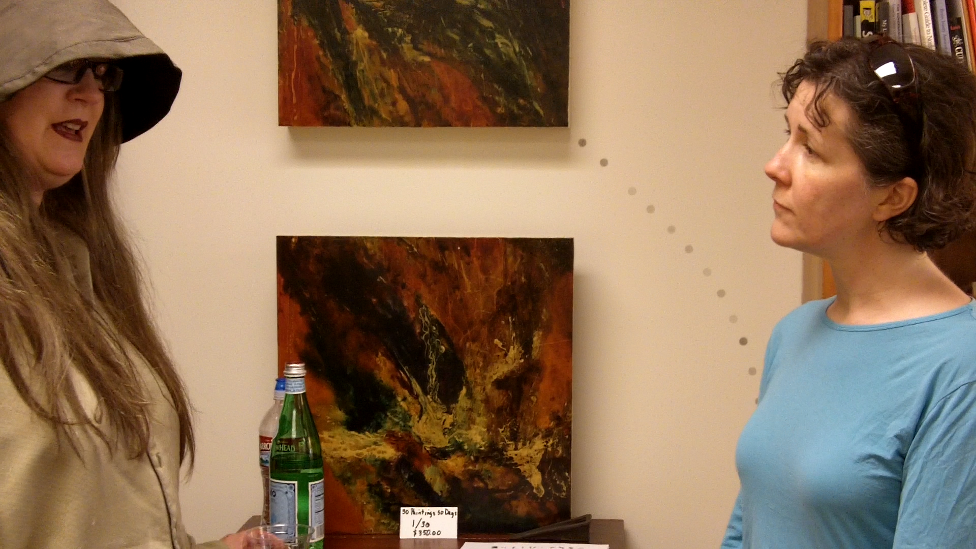 Kris Haas and Lynn Keating talking Two women in profile are talking. Between them are abstract paintings on the wall. One woman has a dark hat pulled down over her eyes.