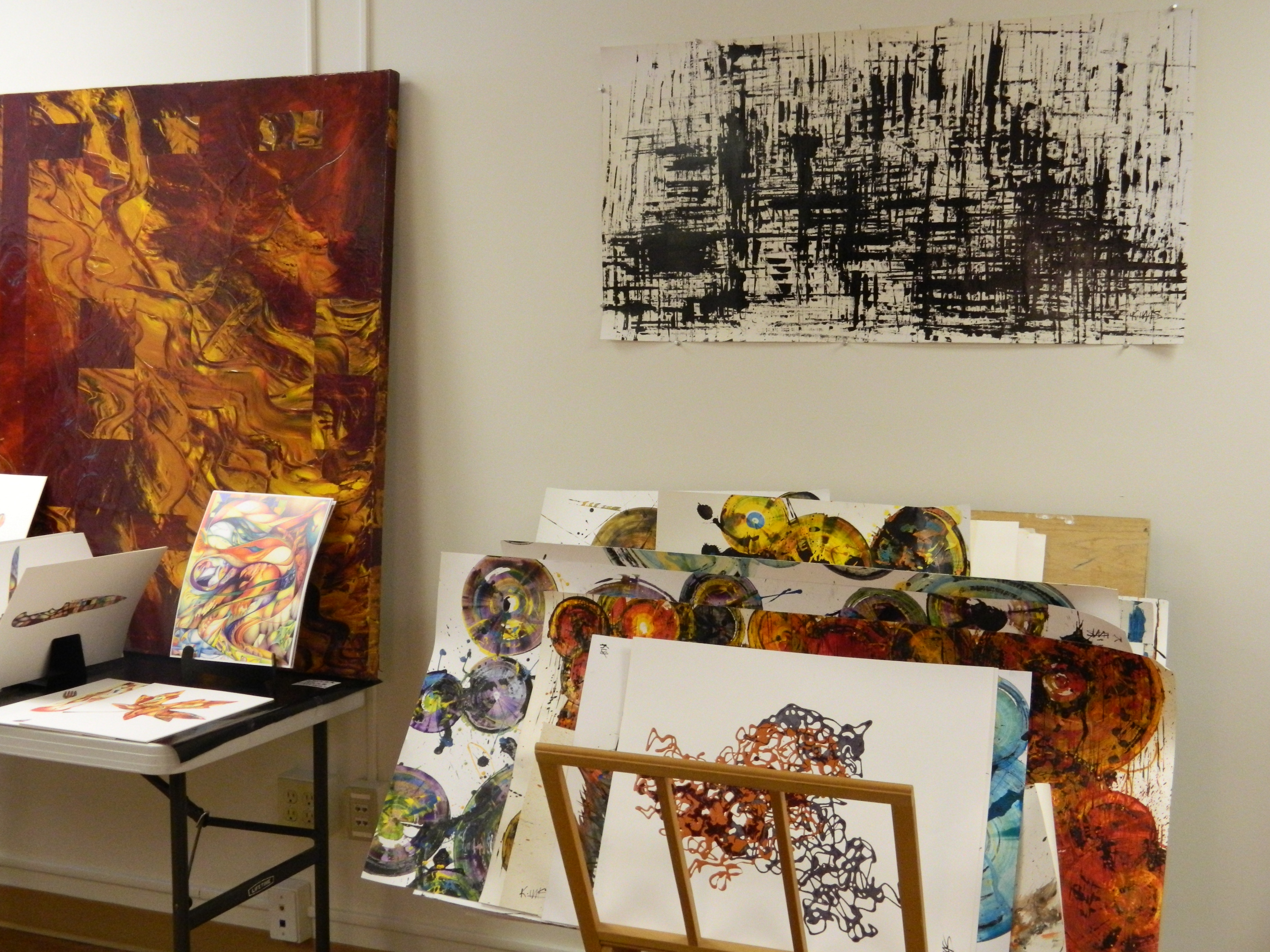 Kris's artwork at the reception Abstract paintings on displayon the wall, resting on a table, or stacked. Images show rich, dark colors, vibrant lines and spheres, and splashes.