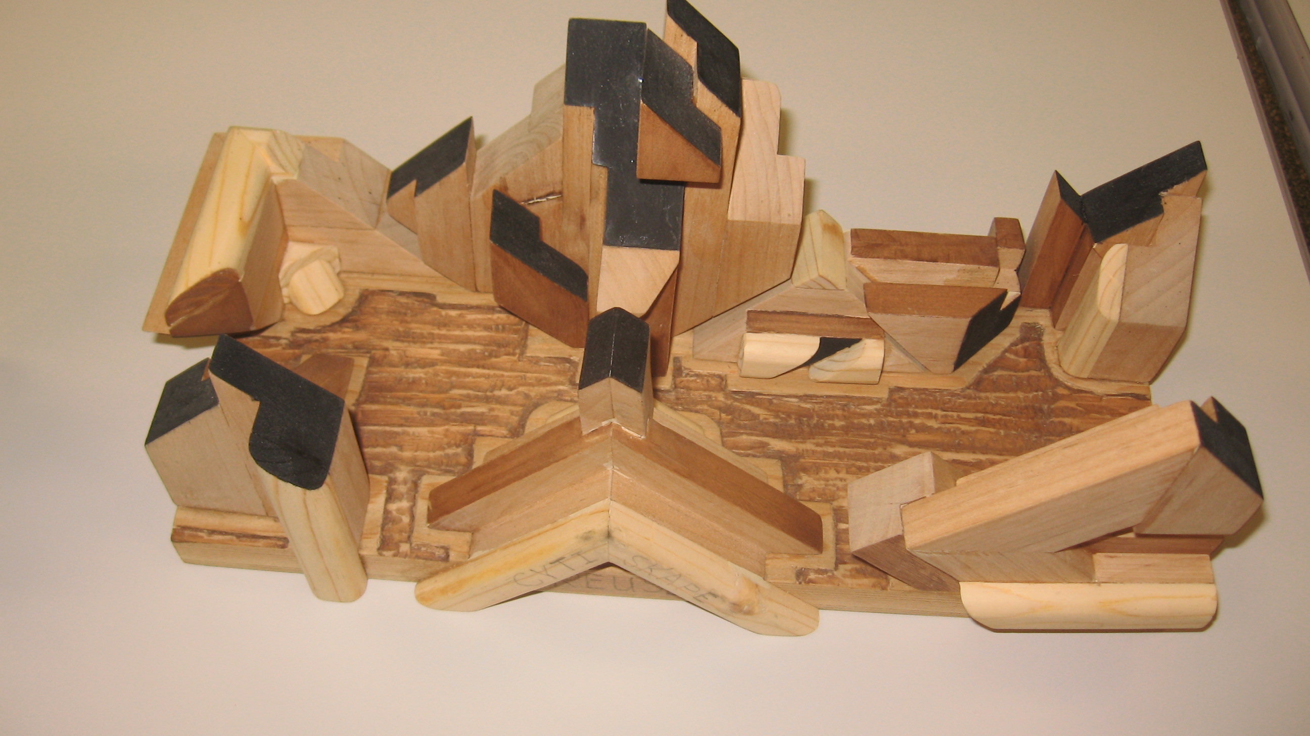 "Cyti Skape" wood carving An intricate three-dimensional wood carving showing a representation of a city scape. The wood is blonde, and tops of buildings are black.