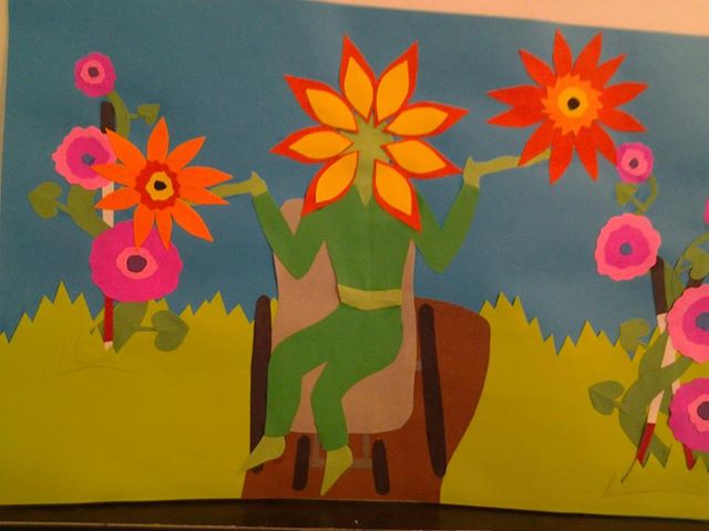A green human figure sits in a brown chair. Its head is a large, vibrant flower, and its hands are open to the sky. All around the figure are other bright pink, yellow, orange, and red flowers. The images are all cut paper, and you can see the layers and texture of the paper.