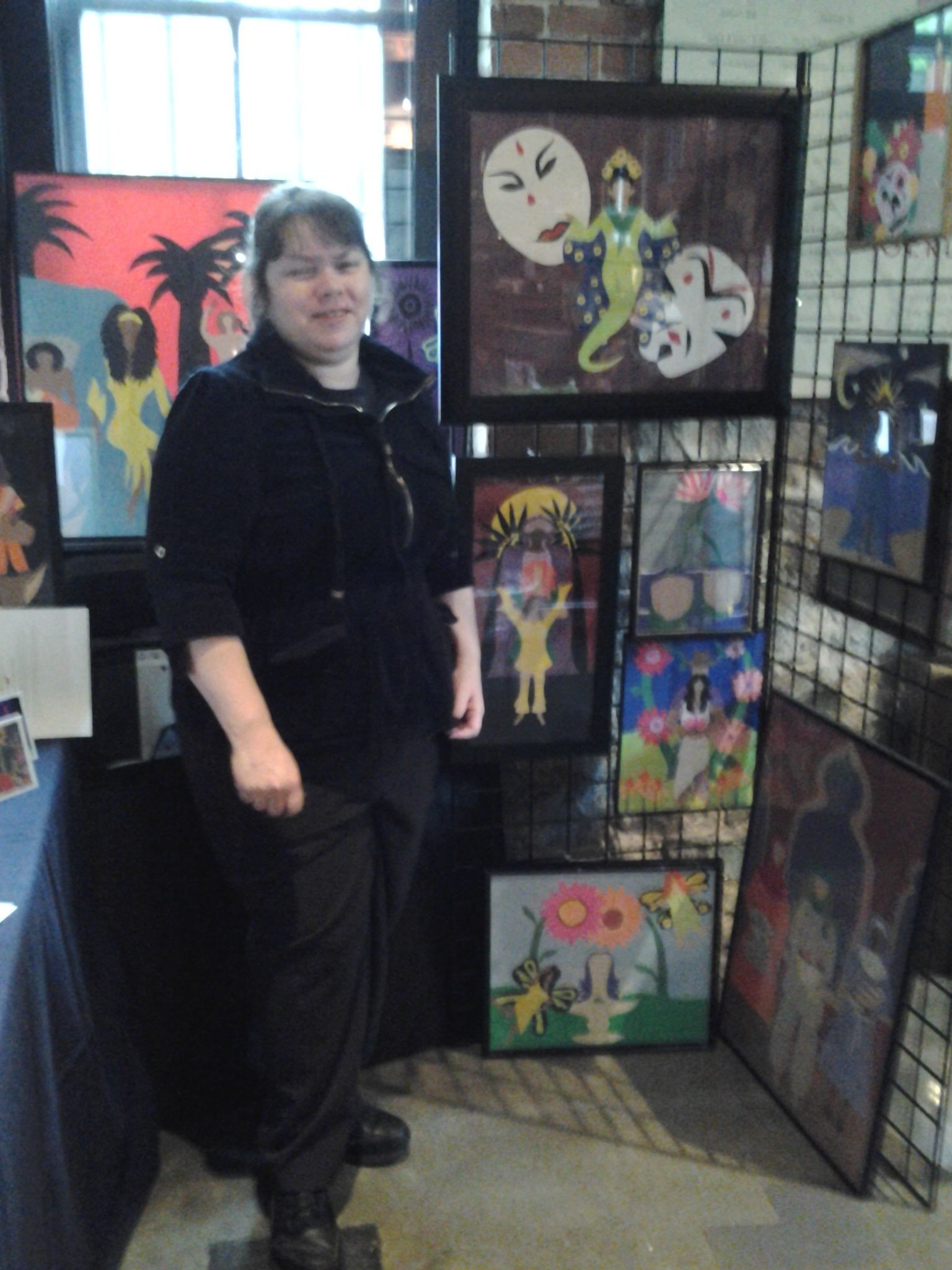 Lavaun Heaster with papercut artwork A woman in black stands next to a metal stand displaying framed artwork. Artwork is multicultural, multi-ethnic visual arts created with many colors of cut paper.