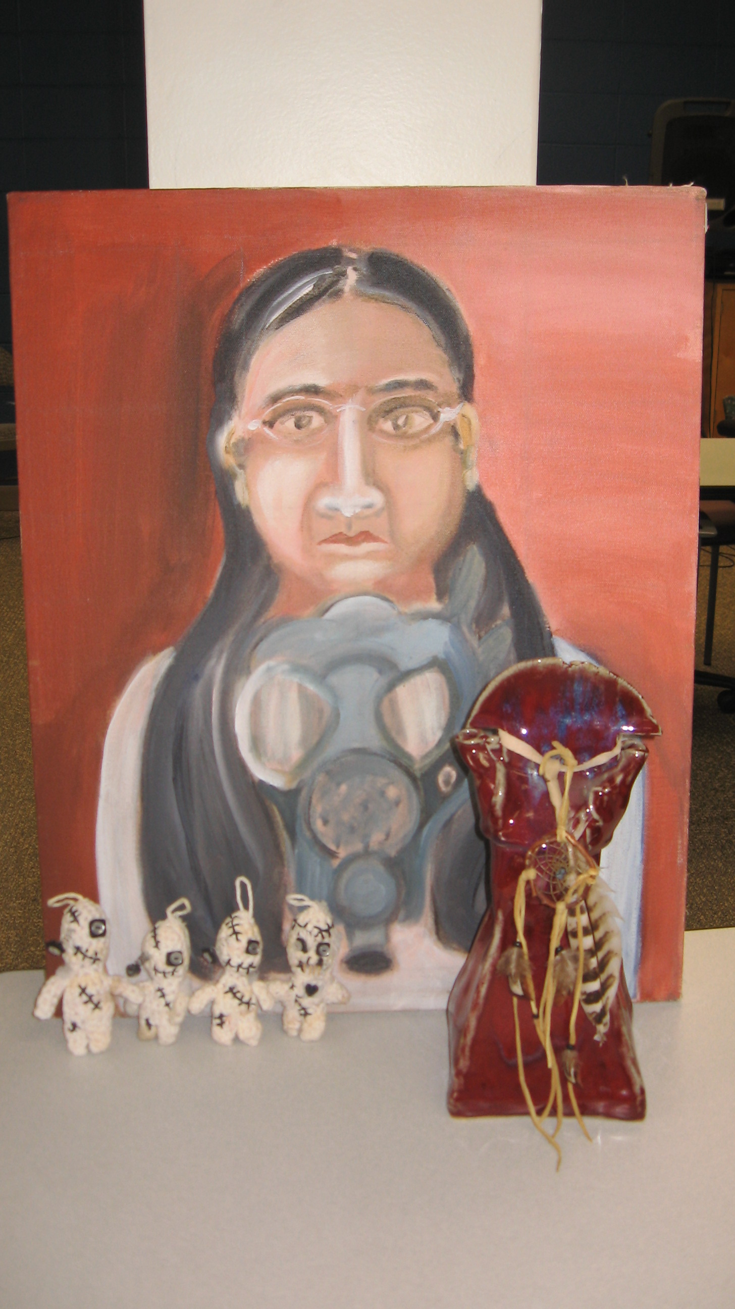 painting and sculptures An earth toned painting of a dark-skinned woman with glasses and long hair holding a gas mask in front of her. She looks solemn. Four small soft sculptures of white knitted yarn that are somewhat human shaped. They are covered in surgical stitches and each have one button eye hanging off. A burgandy glazed ceramic sculpture with a dream catcher and feathers.