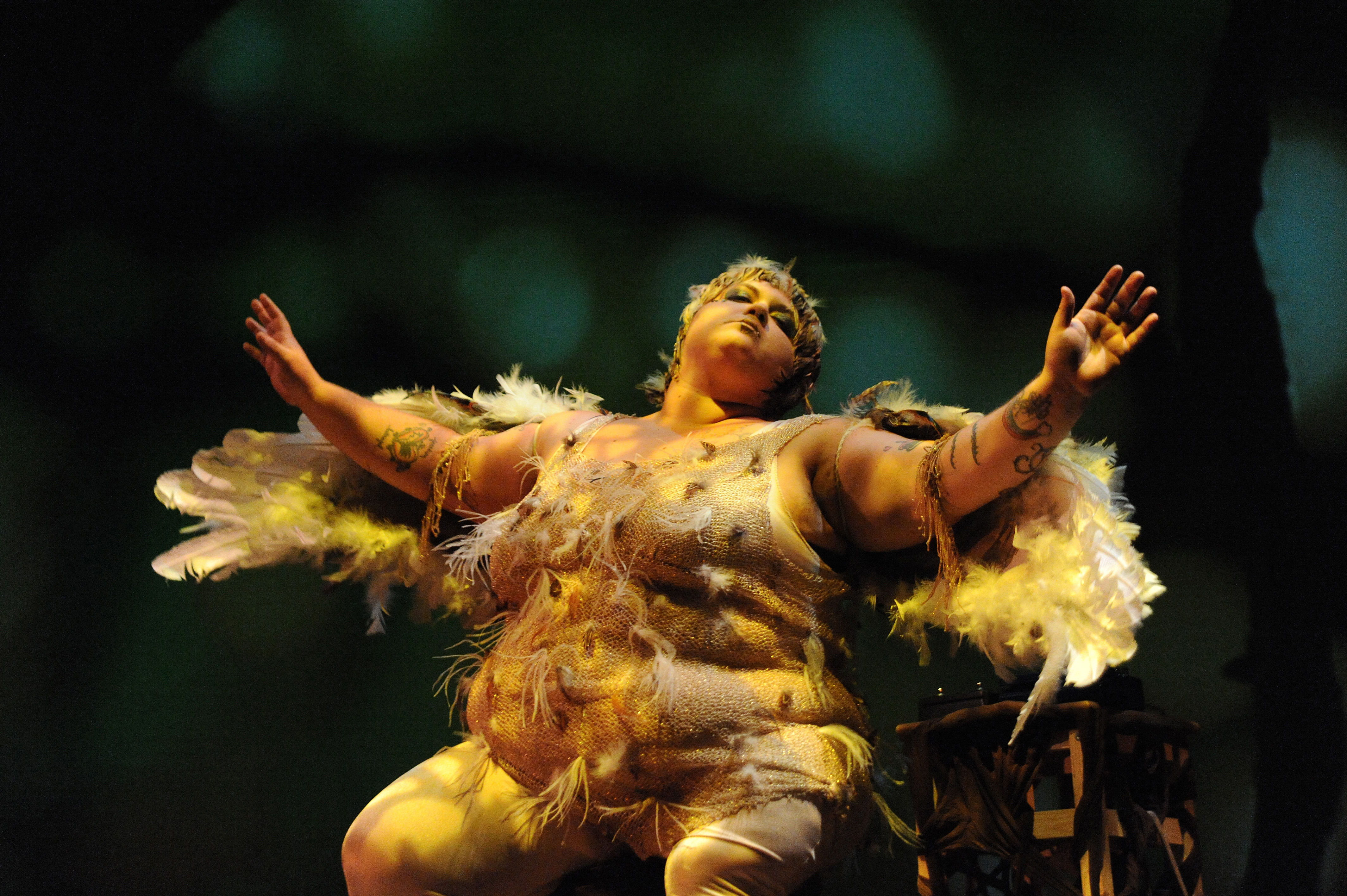 Still image of Nomy Lamm from the documentary A fat woman of color sits perched on a stool with her arms outstretched, her head tilted back, and her eyes almost closed in a blissful, thoughtful look. She is costumed as an angelic looking bird, glittering and feathered. The lights are low and warm, with shadows cast along her body.