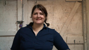 Portrait of Cynthia, a white woman with shoulder-length red hair tied back into ponytails. She stands by a barn door, smiling.