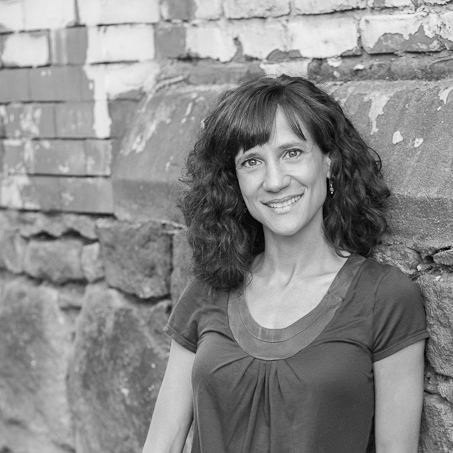 Black and white photo of Cheryl, a white Ashkenazi Jewish woman with olive complexion, shoulder-length, curly dark hair, and gray-green eyes. She stands in front of a rough brick wall and stone wall smiling for the camera.