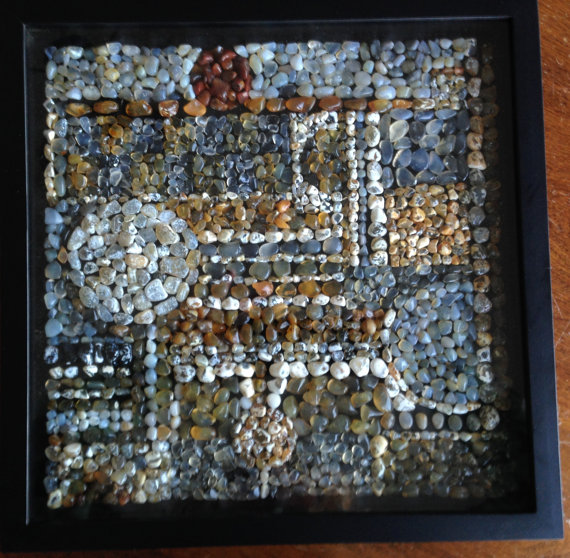 A shadow box with black frame on a dark wood table. Inside the box is an intricate, geometrical design with tiny, densely packed pieces of beach glass. Colors include blues, browns, whites, and yellows. Designs include circles, bars, lines, and stripes.