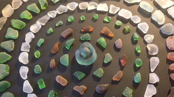 Six rings made of small beach glass pieces are visible, making up a mandala. Each ring is one color family, and the rings are pinks, greens, whites, and reds. In the center is a larger piece of beach glasses. The rings are on rich brown, textured paper.