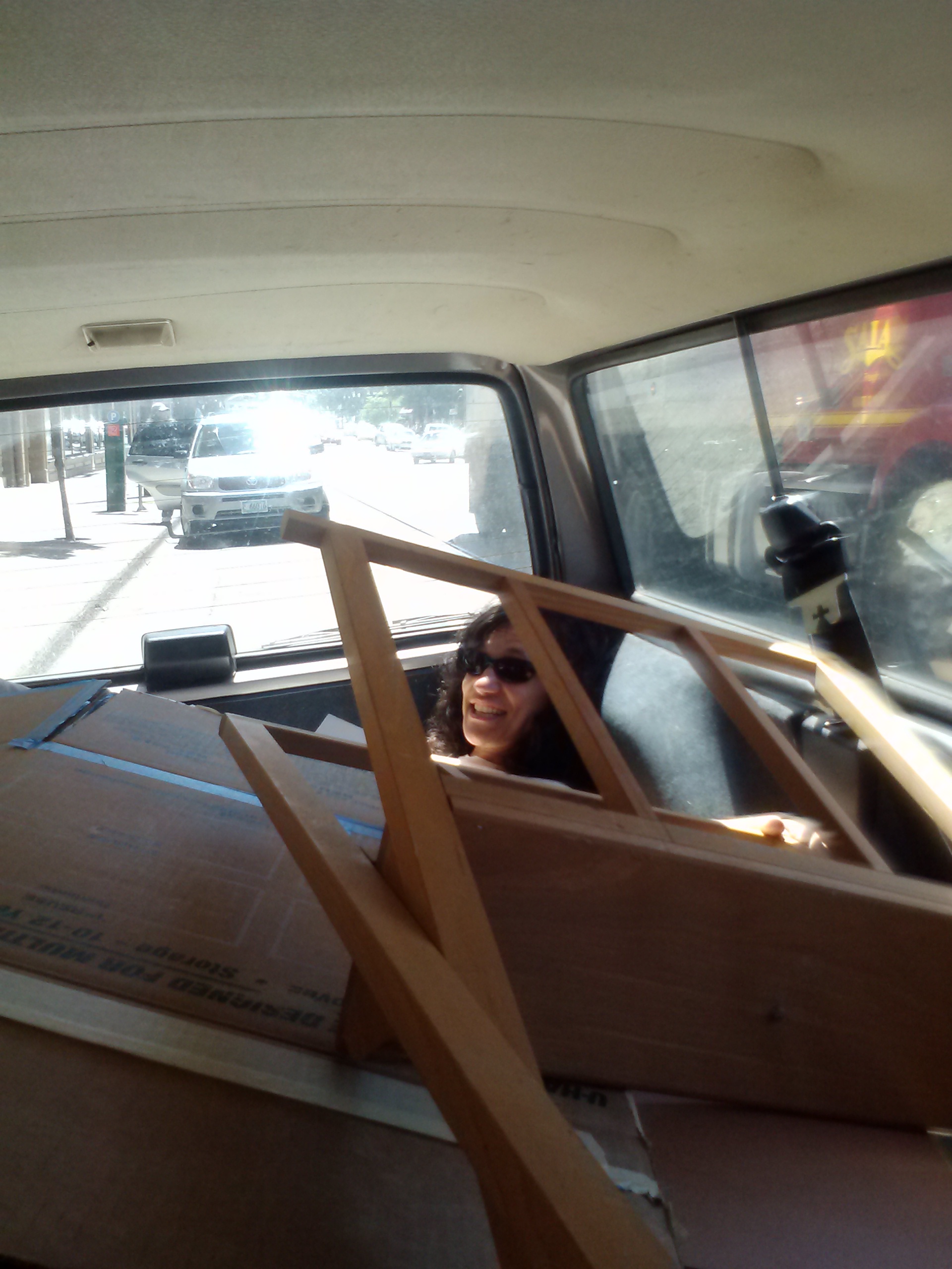 Cheryl, wearing sunglasses, is lying in the back of a station wagon trunk underneath a large pile of paintings. 