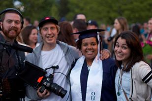 Four people smile for the camera: Shawn and Paulius hold their recording equipment, Dani is in blue and white high school graduation cap and gown, and Athena is in a striped sweater.