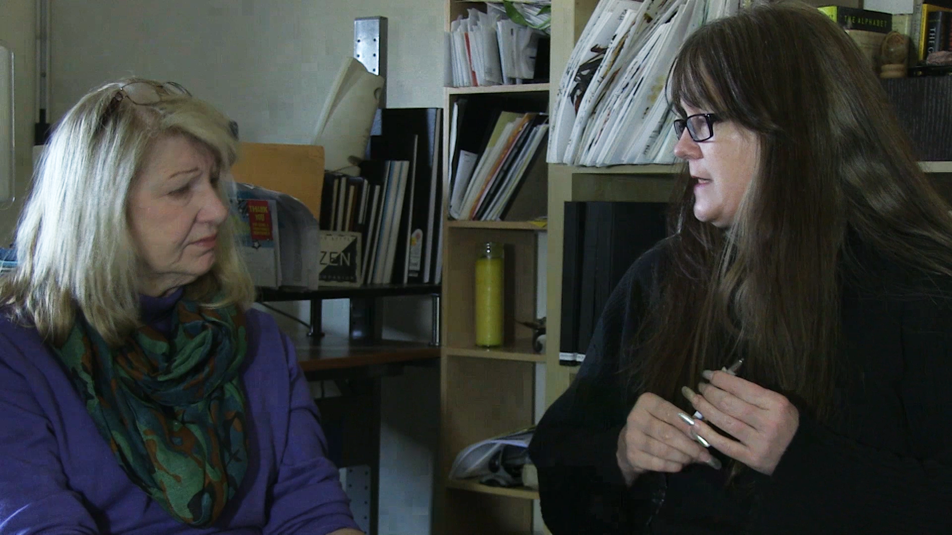 Kris and Pat sit next to each other, talking, with stacks of paintings on the shelves behind them in Kris's studio.