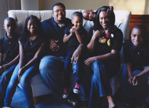A professional family portrait of Morénike Giwa Onaiwu, her husband, and  their children. They all wear dark t-shirts and blue jeans. Smiles range from serene to exuberant.