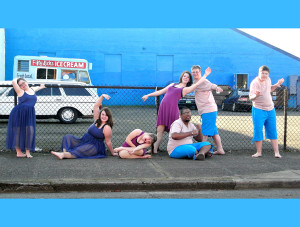 Seven teen and young adult dancers strike poses in front of a bright blue building.
