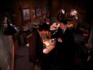 At the police station, Truman stands near the chalkboard with clues. Cooper holds up the jar of burnt engine oil for Truman to smell. The Log Lady, Pete, and Hawk watch them from across the room.