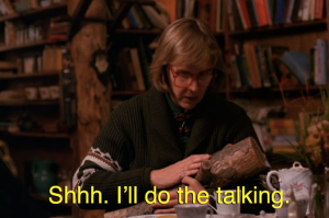 The Log Lady sits at her dining room table in a room filled with books, looks down at her log lovingly, and strokes it reassuringly