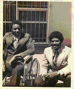 Old photo of two smiling Black men in suits: Brad Lomax in a wheelchair and Glen Lomax, crouching down.
