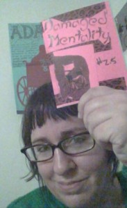 A woman with glasses holds up a small, pink zine "Damaged Mentality #2.5"