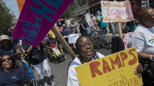Photo of NYC Disability Pride parade showing marchers in wheelchairs and on foot. Main person in the photo is a Black woman holding protest sign.