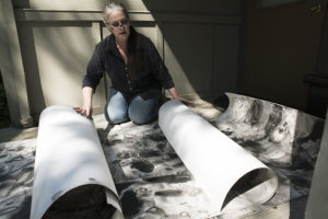 Kris Haas kneels on the ground, unrolling 18-foot sheets of paper with black and gray ink paintings.