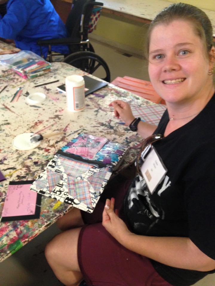 A woman smiles for the camera and shows off her colorful collage project, a handmade journal.