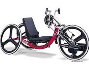 A red three-wheeled cycle, low to the ground. Pedals are attached to the front wheel and are operated by hand.