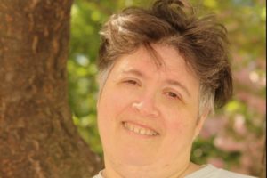 A white woman with short, graying hair stands outside on a sunny day near trees and smiles.