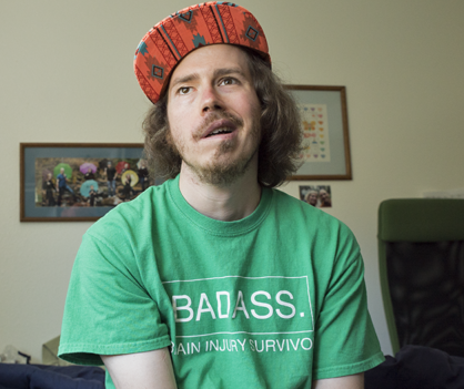 Brandon Scarth smiles. A portrait of his family is on the wall behind him. He wears a colorful cap and a green t-shirt with "BADASS Brain Injury Survivor" in white lettering. Copyright 2016 Sika Stanton.