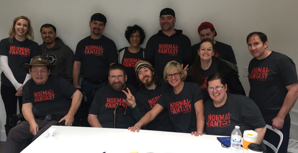 13 people smiling for the camera. 12 are wearing matching shirts with "Normal Is A Fantasy" in red letters. Most people in the photo have a TBI or an ABI, and some have other disabilities too.