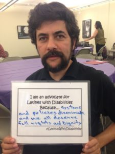 A Latinx person with dark wavy hair and beard holds a sign. "I am an advocate for Latinos with Disabilities because...systems and policies discriminate and we all deserve full rights and dignity. #LatinoswithDisabilities