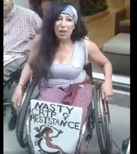 Maria Palacios, a Latina in a tank top and skirt, sits in a manual wheelchair. Resting on her legs is a sign: "Nasty Crip Resistance. Free our people." There is a Venus woman power symbol and a nude Brown woman in a wheelchair, arms raised, chains around her wrists broken, adapted from the ADAPT logo.