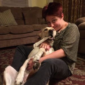 Colleen Connor, a white woman with short magenta hair sits on the living room floor. A golden dog lies on its back, cradled in Colleen's lap. A large white bone dangles out of the dog's mouth, and Colleen's hand is on the dog's belly.