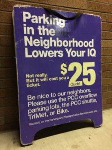 A purple Portland Community College sandwich board sign with white and yellow lettering leans against a brick wall. It reads: "Parking in the neighborhood lowers your IQ. Not really. But it will cost you a ticket. $25. Ouch. Be nice to our neighbors. Please use the PCC overflow parking lots, the PCC shuttle, TriMet, or Bike. Find info on the Parking and Transportation Services web site."