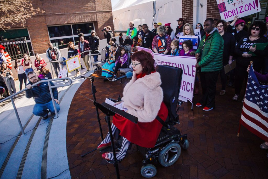 Caitlin Wood, a white woman in bright red dress, fuzzy coat, and striped stockings sits in a power wheelchair and addresses a crowd. Behind her stand and sit people of various ages, ethnicities, and genders holding Fayetteville Women's March signs.