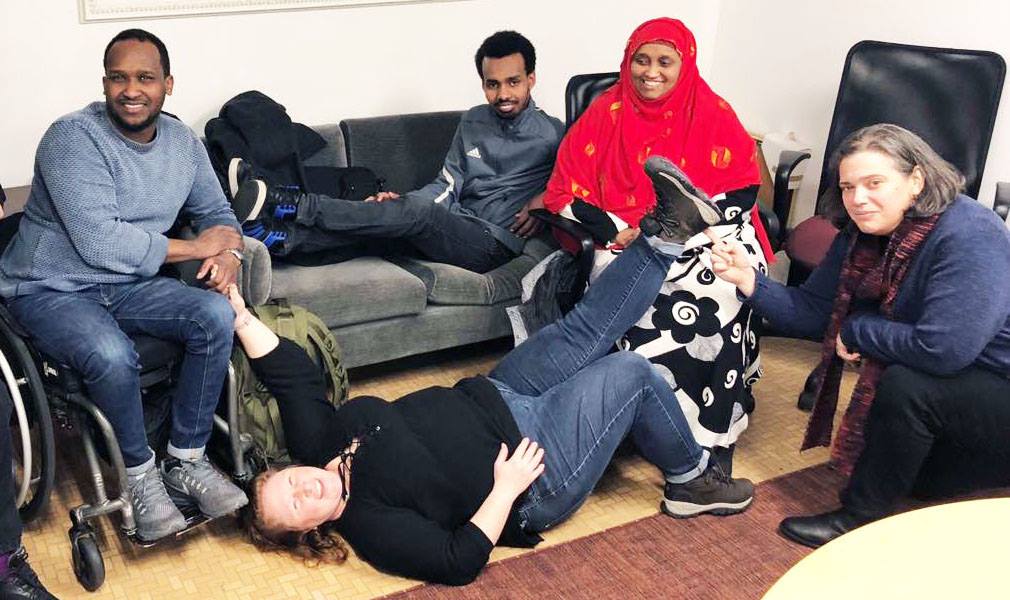 Photo by Shannon Sullivan. Image description: Five people sit on a chair and in a wheelchair, lay on a couch and the floor, and kneel on the floor, smiling and striking poses.