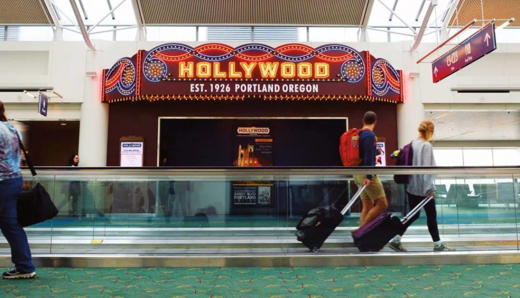 Hollywood Theatre microcinema at the airport. People walking past with their luggage. The entry is an ornate, old-fashioned theater design with lights. It says "Hollywood. Established 1926, Portland, Oregon."