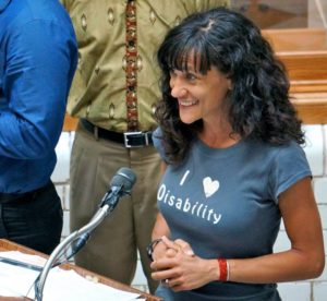 Me, a white woman with olive complexion and long, curly brown hair, speaking into a microphone on a podium. I'm clasping my hands tightly and smiling tightly. My shirt has iron-on I (heart) disability. The heart is a brain designed in the shape of a heart.