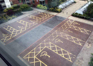 A view from my dorm room window at Liverpool Hope University shows a parking lot with only eight slots. All eight are designated accessible, and they all have yellow lines painted between them to leave space for accessible van ramps. Then, two rows of bike racks with roll-down rain and snow covers.