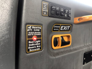 The inside of a Manchester taxi door. A bright yellow grab bar. Some switches. A sign with "Hearing Loop. Switch hearing aid to t-position." A lit red light and "Red light indicates doors are secured."