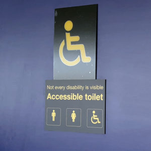 Signs on the bathroom door in England airport. A very large image of the universal access logo of the stick figure in a wheelchair. Below that, "Not every disability is visible. Accessible toilet." Then, the standard images showing male, female, and disabled user.