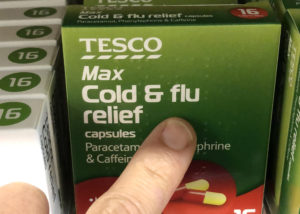 My finger touching the box of a Tesco brand cold and flu relief medicine. Text on the box lists the active ingredients in white lettering and in Braille.