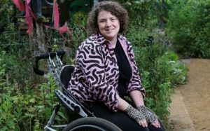 Jess, a white woman with curly brown hair, sits outside in a custom manual wheelchair. She wears a pink jacket with black squiggles and leopard print fingerless gloves and smiles for the camera.